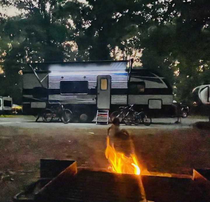 Enjoying an RV rental at night. A brightly lit travel trailer with its awning out, parked among trees, campfire glowing foreground.. Heartland Prowler 2021