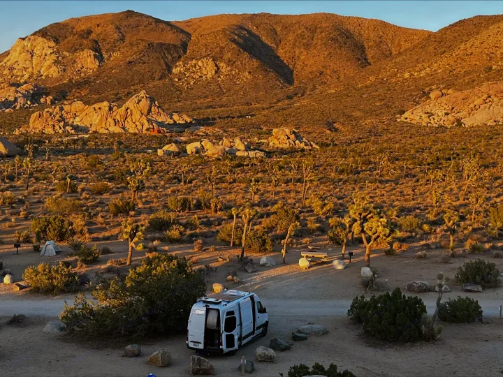 RV rental parked at a desert campsite among Joshua trees and mountains at golden hour. Perfect for a nature retreat.. Mercedes-Benz Sprinter 2021