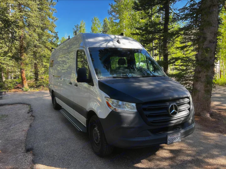 A white Mercedes Sprinter camper van (RV rental) in a sunny, wooded campsite. Perfect for nature road trips & adventure.. Mercedes-Benz Sprinter 2021