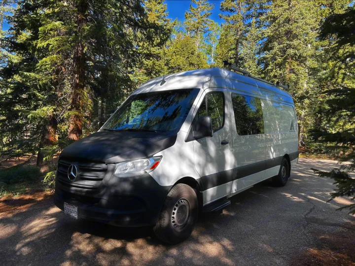 A white Mercedes-Benz Sprinter van, an RV rental, with a black hood, parked on asphalt amidst tall green pine trees.. Mercedes-Benz Sprinter 2021