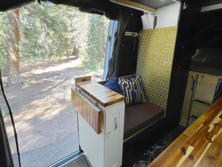 RV rental camper van interior. Cozy space with a pull-out counter, golden hex wall, and forest view from the open door.. Mercedes-Benz Sprinter 2021