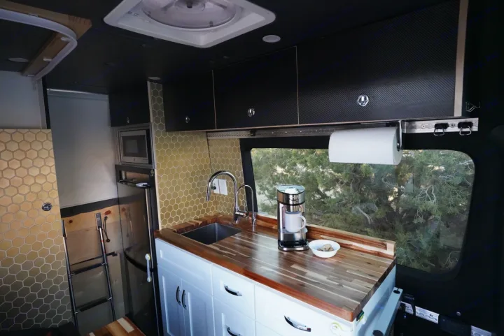 RV rental interior: Custom kitchen with wood counters, sink, microwave, and hex-tile backsplash. Trees visible outside.. Mercedes-Benz Sprinter 2021