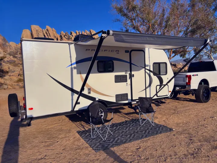 RV rental ready: White travel trailer with extended awning, chairs, and mat on a dirt lot, hitched to a truck, amidst desert rocks.. Pacific Coachworks Pacifica XL 2020