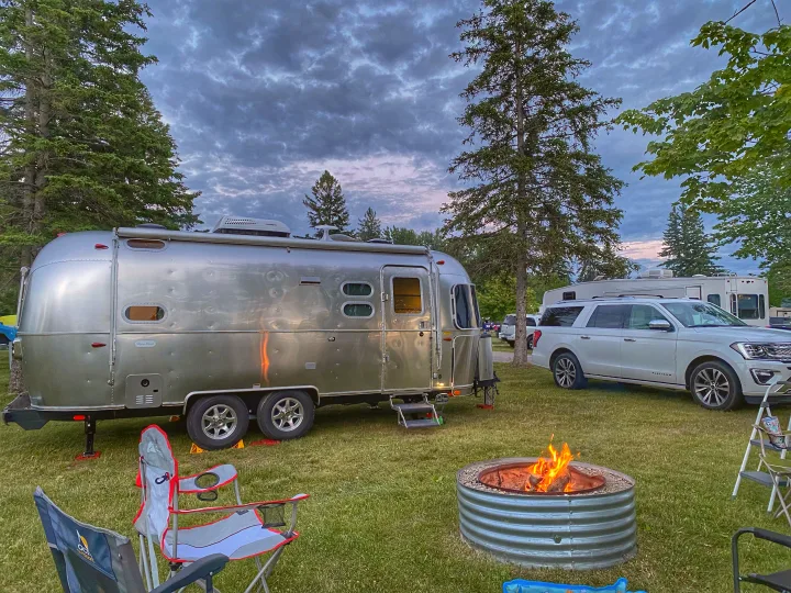 A silver Airstream rv rental and white SUV at a twilight campsite with a glowing fire pit and chairs.. Airstream Flying Cloud 2022