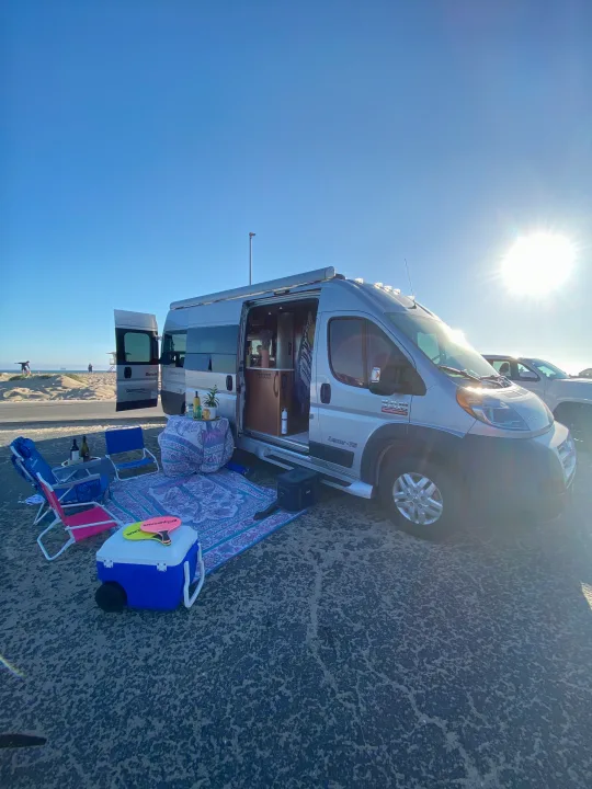 Perfect beach setup with an RV rental! Silver camper van, purple rug, chairs, and cooler on asphalt by sandy dunes & ocean under bright sun.. Pleasure-Way Pleasure-Way Class B 2016