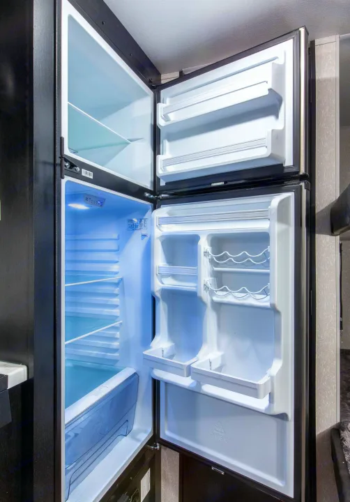 Open two-door RV rental refrigerator & freezer. Brightly lit interior with empty shelves and door storage, including bottle racks.. Forest River Cherokee Wolf Pup 2022