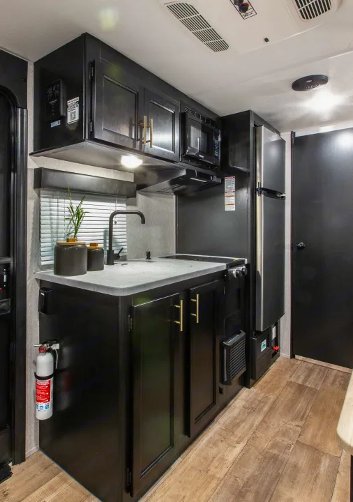 Inside this RV rental, a modern black kitchen with a sink, microwave, fridge, stovetop, and gold-handled cabinets.. Forest River Cherokee Wolf Pup 2022