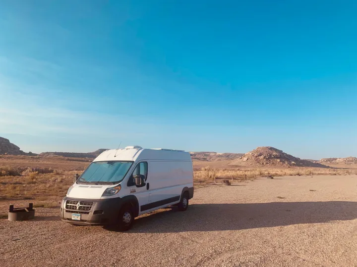A white Ram ProMaster RV rental campervan is parked in a desert campsite with rocky hills under a blue sky.. Other Sprinter Van 2017