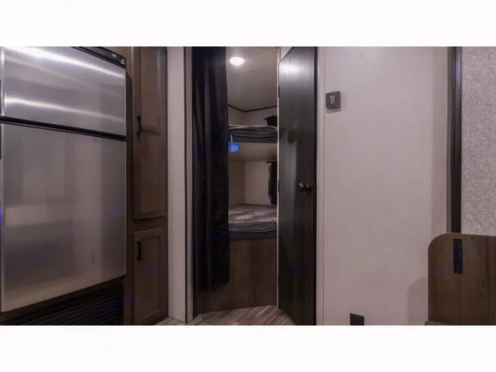 Compact RV rental interior featuring a stainless steel fridge, adjacent cabinets, and a bunk bed area with a privacy curtain.. Autumn Ridge 26BHS 2022
