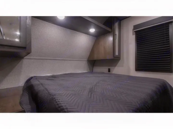 Compact RV rental bedroom featuring a dark gray bed, built-in overhead storage with lights, and a window with blinds.. Autumn Ridge 26BHS 2022