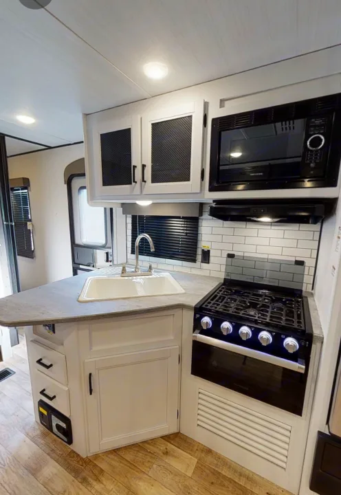 Bright RV rental kitchen with white cabinets, sink, and black appliances: microwave, range, and a subway tile backsplash.. Keystone Hideout 2022