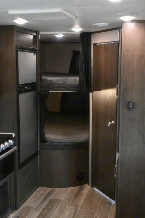 Inside an RV rental: bunk beds with privacy curtains, a refrigerator, and a kitchen area, all with dark wood finishes and light flooring.. Jayco Jay Flight SLX 2021