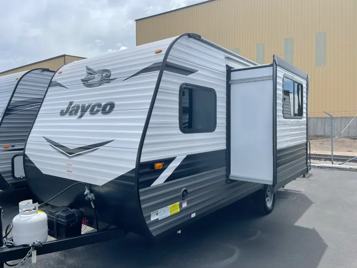 Jayco travel trailer for RV rental, showcasing its side with an open utility door, window, and front propane tank.. Jayco Jay Flight SLX 7 184BS 2022