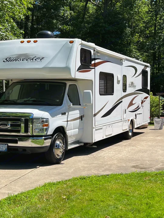 This white Sunseeker RV, perfect for your next rv rental adventure, is parked on a sunny driveway surrounded by trees.. Forest River Sunseeker 2013