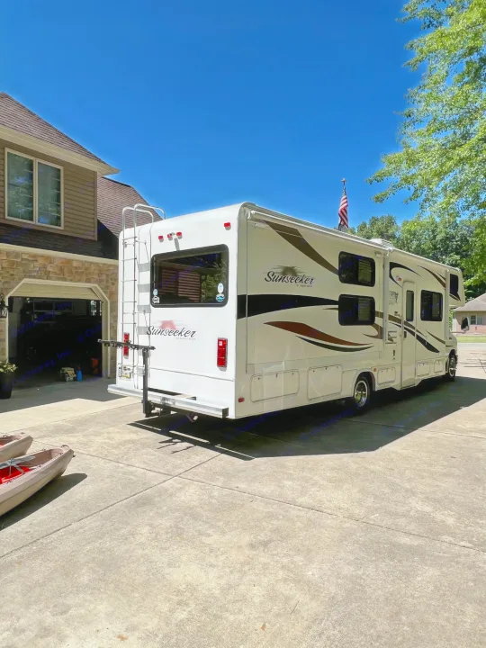A white Sunseeker RV with a US flag, parked on a sunny driveway. Consider this RV rental for your next adventure.. Forest River Sunseeker 2013