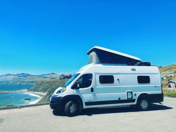Your next RV rental awaits! Enjoy coastal views from this white pop-top camper van, parked beside a bright blue ocean.. Winnebago solis 2022