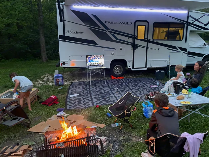 Family enjoying an RV rental campsite. Outdoor TV, glowing campfire, and dining create a cozy evening.. Other Other 2022