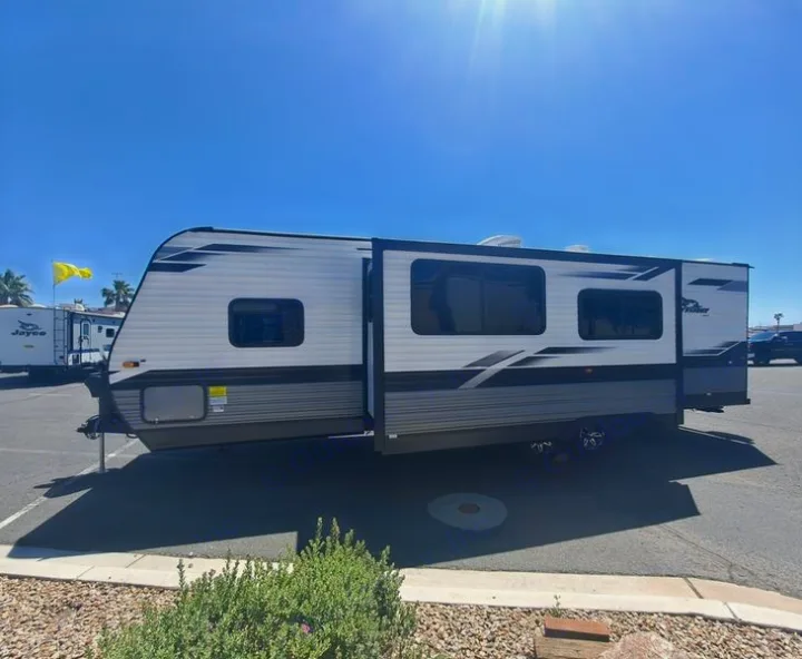 White & gray travel trailer, an RV rental, with a slide-out, parked on a sunny day in a paved lot.. Jayco JayFlight 28BHS TT 2022