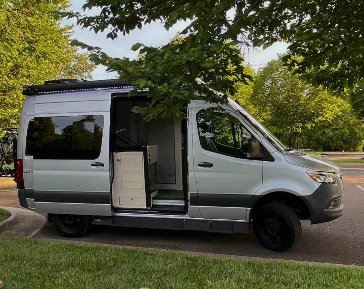 A sleek silver camper van, perfect for rv rental, is parked among green trees with its side door open, revealing the interior.. Tiffin Motorhomes Cahaba 2021
