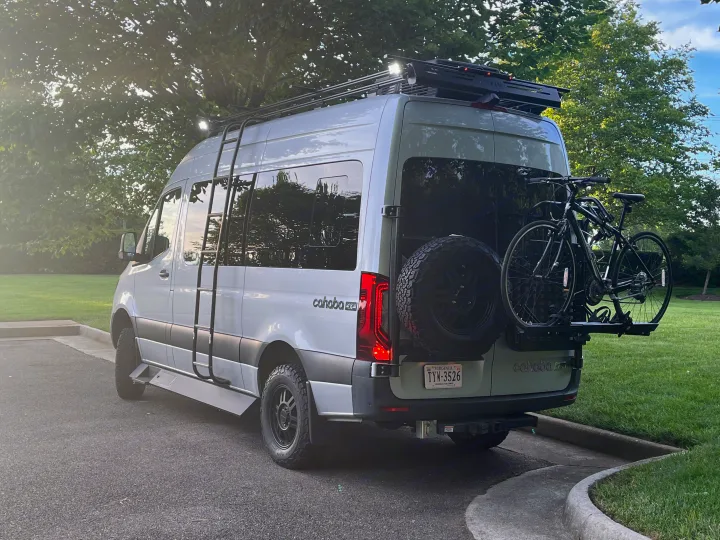 Light gray Cahaba 4x4 camper van, ideal for RV rental, with a roof rack, side ladder, rear spare tire, and two bikes mounted.. Tiffin Motorhomes Cahaba 2021