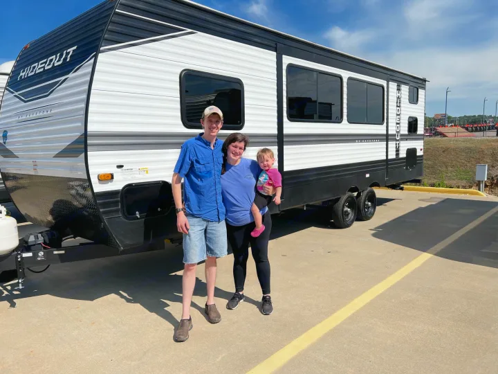 Proud family (man, woman, toddler) smile in front of a white and black Hideout RV. Ideal for your next rv rental journey.. Keystone Hideout 2022