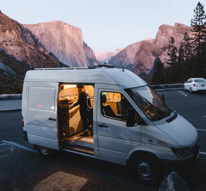 Person inside a cozy, illuminated RV rental camper van, parked amidst iconic Yosemite Valley mountains at dusk.. Mercedes Sprinter 2006