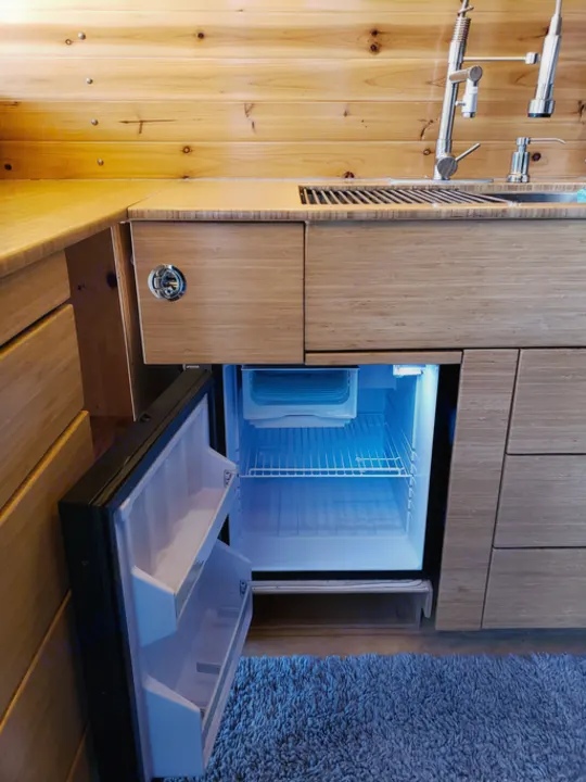 Modern RV rental kitchen featuring a compact fridge with a glowing blue interior, wood cabinetry, sink, and rug.. Mercedes Sprinter 2006