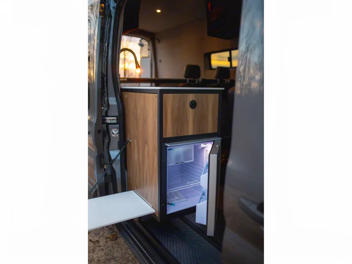 RV rental interior: a compact kitchen with wood cabinets, a lit fridge, and a pull-out table.. Ford Transit 2021