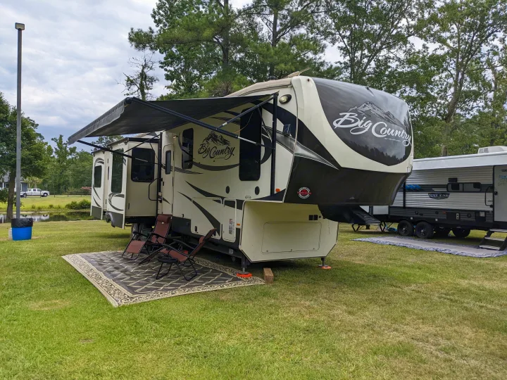 A spacious fifth-wheel RV rental, featuring an extended awning, outdoor rug, and chairs, set up on a grassy campsite.. Heartland Big Country 2016