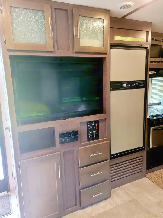 Inside an RV rental: a modern entertainment center with TV, Furrion stereo, controls, fridge, and stylish wood cabinetry.. Crossroads RV Sunset Trail Super Lite 2018
