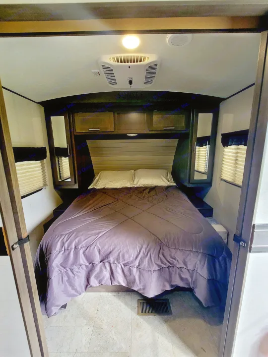 This RV rental bedroom interior features a cozy bed with purple bedding, overhead storage, an AC unit, and windows.. Crossroads RV Sunset Trail Super Lite 2018