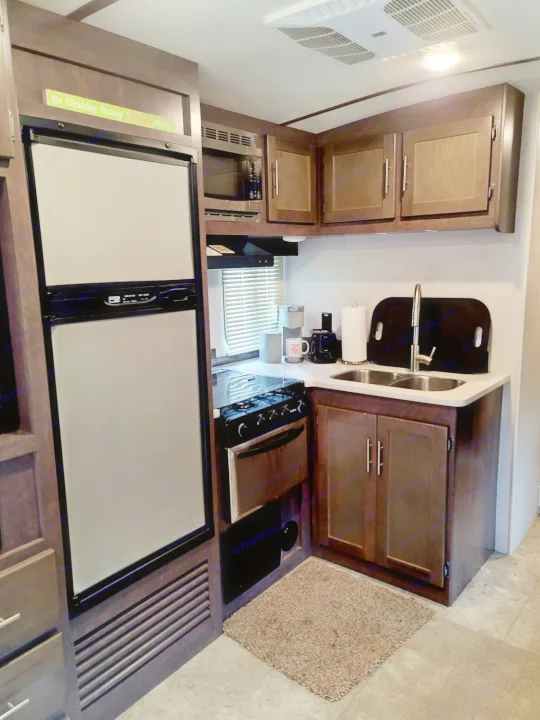 Compact RV rental kitchen featuring a fridge, microwave, stove, sink, and a fun "No Working During Drinking Hours" sign.. Crossroads RV Sunset Trail Super Lite 2018