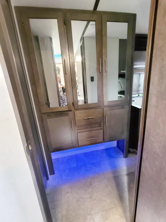 RV rental interior: Wooden cabinet unit with mirrored doors reflecting a person and sink, storage drawers, and blue floor lighting.. Crossroads RV Sunset Trail Super Lite 2018
