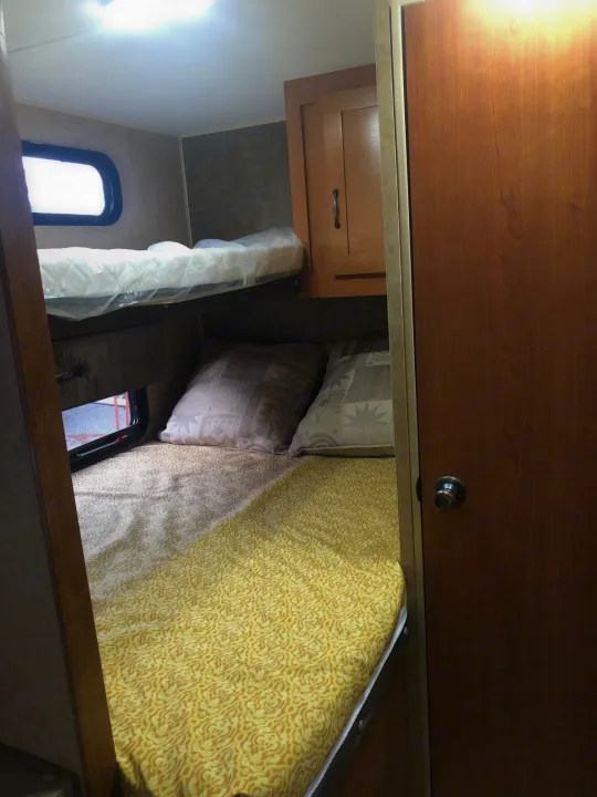 Compact RV rental bedroom showing a main bed with yellow patterned bedding, pillows, and an upper bunk bed with new mattress.. Forest River Cherokee 2015