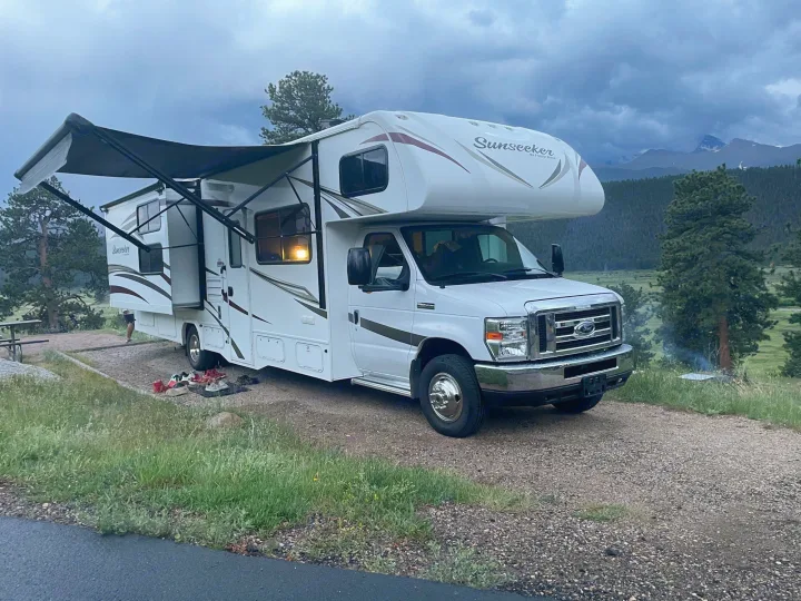 A white rv rental with its awning and slide-out extended at a scenic campsite among trees, mountains, and clouds.. Forest River Sunseeker 2017