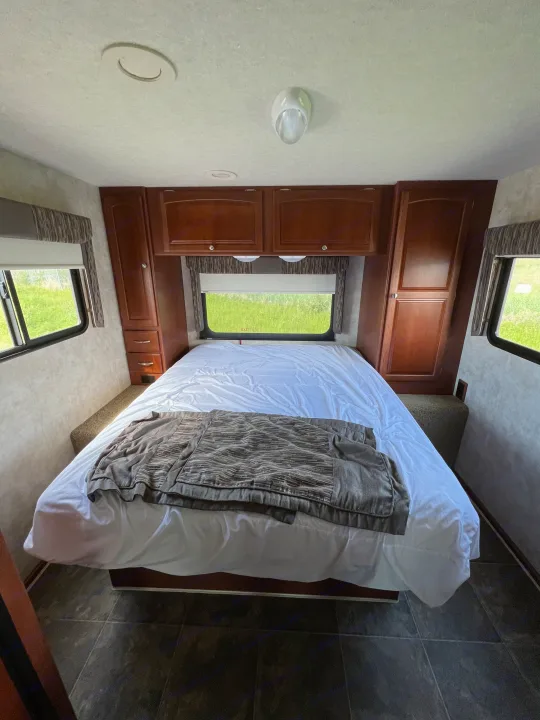 Interior of an RV rental bedroom, featuring a large bed, built-in storage, and bright windows with green views.. Winnebago Chalet 2013
