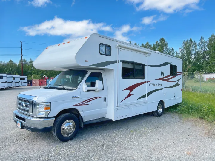 This white Class C RV rental, a Ford E-450 motorhome, is parked on a gravel lot under a bright blue sky.. Winnebago Chalet 2013