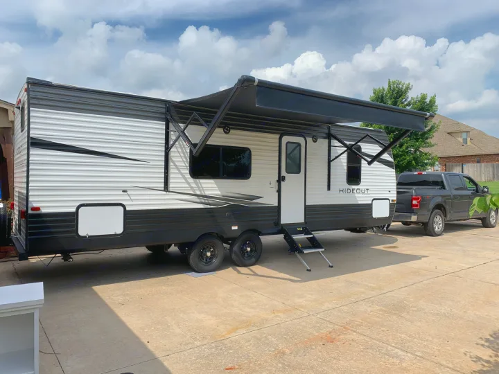 A spacious travel trailer with an extended awning, hitched to a pickup truck on a concrete driveway, ready for an RV rental adventure.. Keystone RV Hideout 2021