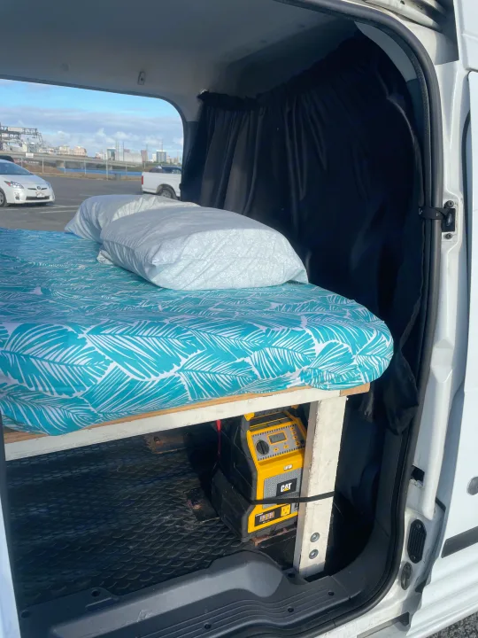RV rental interior: a bed with tropical sheets and pillows, black curtains, and a yellow CAT portable power station inside a camper van.. Ford Transit Connect 2010