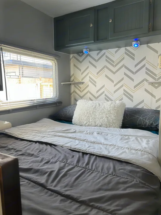 An RV rental bedroom showcasing a bed with gray bedding, dark cabinets, blue lights, and a gray/beige chevron wall.. Dutchmen Sport 2004