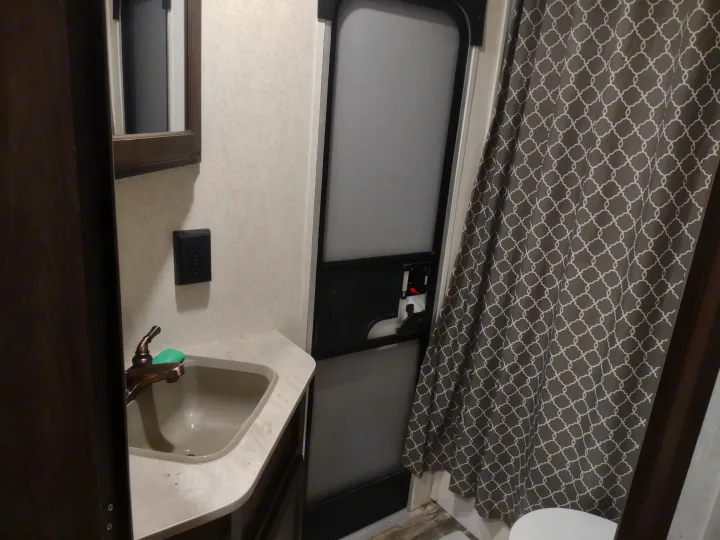 Compact RV rental bathroom featuring sink, faucet, mirror, door, and patterned shower curtain over toilet.. Starcraft Autumn Ridge Outfitter 2019