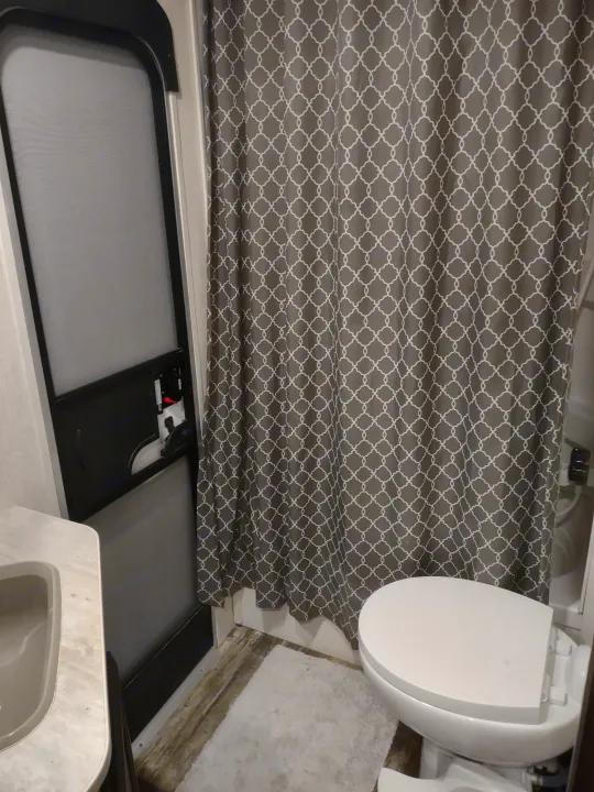 Clean RV rental bathroom featuring a grey patterned shower curtain, white toilet, sink, and wood-look flooring.. Starcraft Autumn Ridge Outfitter 2019