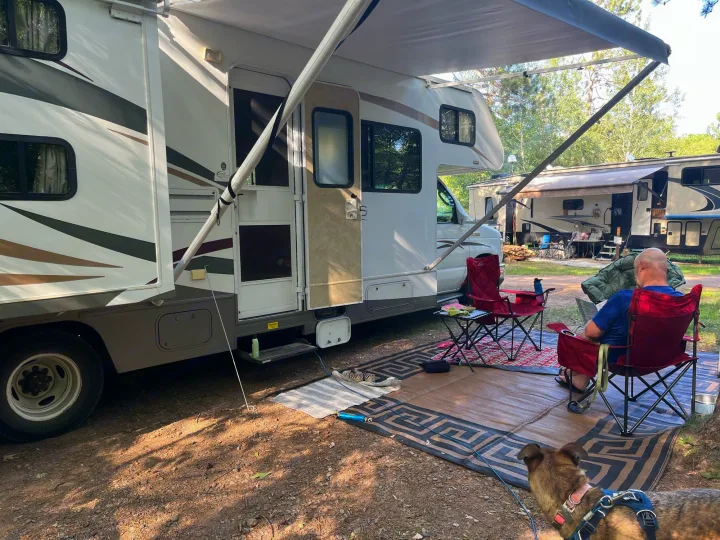 Relaxing at a wooded campsite with an RV rental, an extended awning, a man in a red chair, and a dog.. Winnebago Access 2008