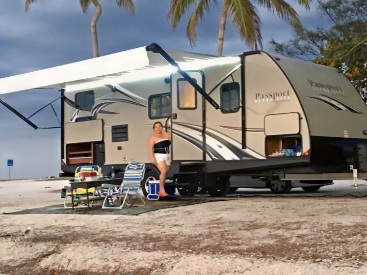 Woman beside her RV rental on a sandy beach. Awning extended for a cozy outdoor camping setup under palm trees.. Keystone RV Passport Elite 2017