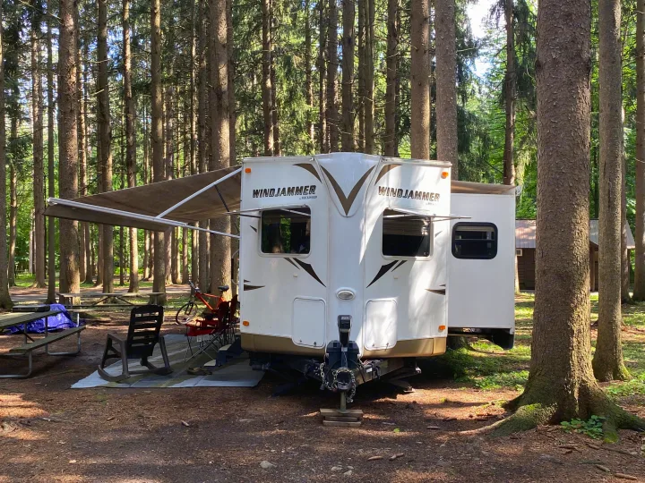 RV rental set up in a serene, shaded forest campground with an extended awning, picnic table, and chairs.. Forest River Rockwood Windjammer 2012