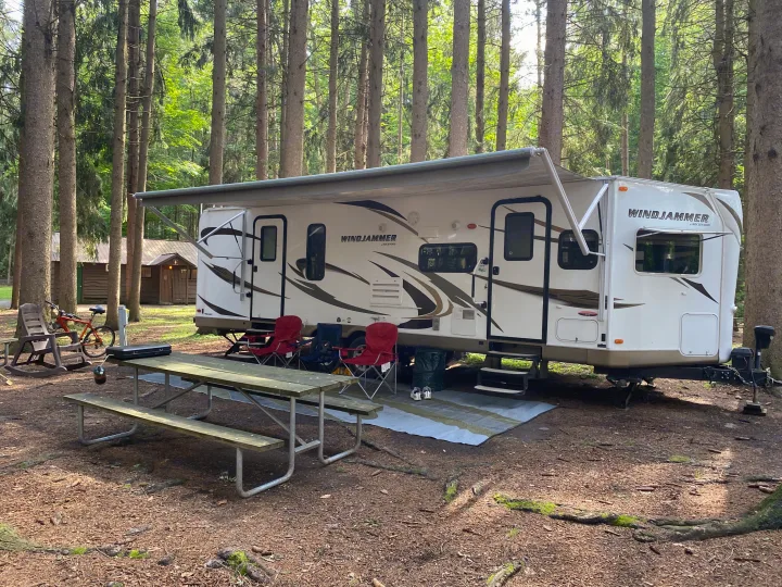 An RV rental is set up under tall trees in a forest campsite, featuring a picnic table, chairs, and a nearby building.. Forest River Rockwood Windjammer 2012