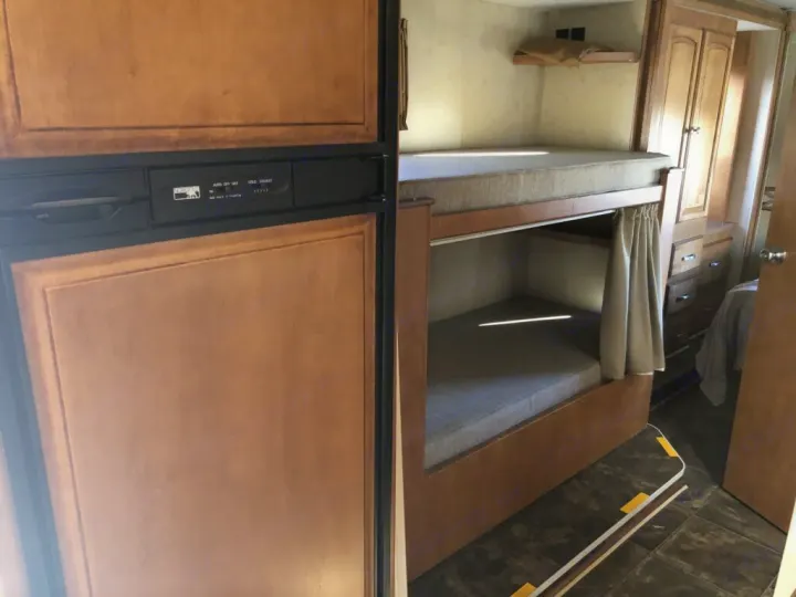 RV rental interior showing a wood-paneled refrigerator, bunk beds with mattresses, and wooden cabinetry. A compact travel space.. Winnebago Chalet 2014