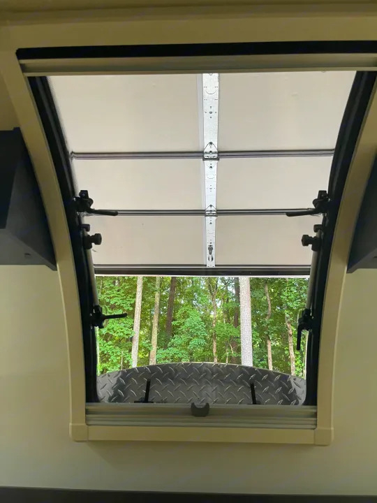 Inside an RV rental, an open window with a pull-down shade reveals lush green trees and a diamond plate surface.. nuCamp RV T@G XL 6 wide 2022