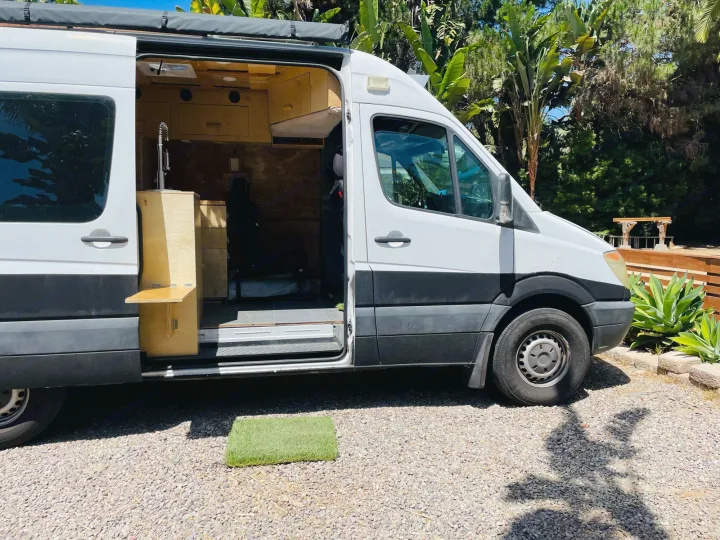 White camper van with custom wood interior, sink, and open door on gravel. Ideal for an RV rental adventure.. Dodge Sprinter Van 2007