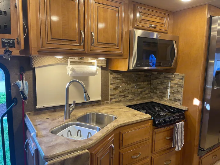 A modern RV rental kitchen featuring light wood cabinets, stainless steel appliances, a sink, and tiled backsplash.. Thor Challenger 2017